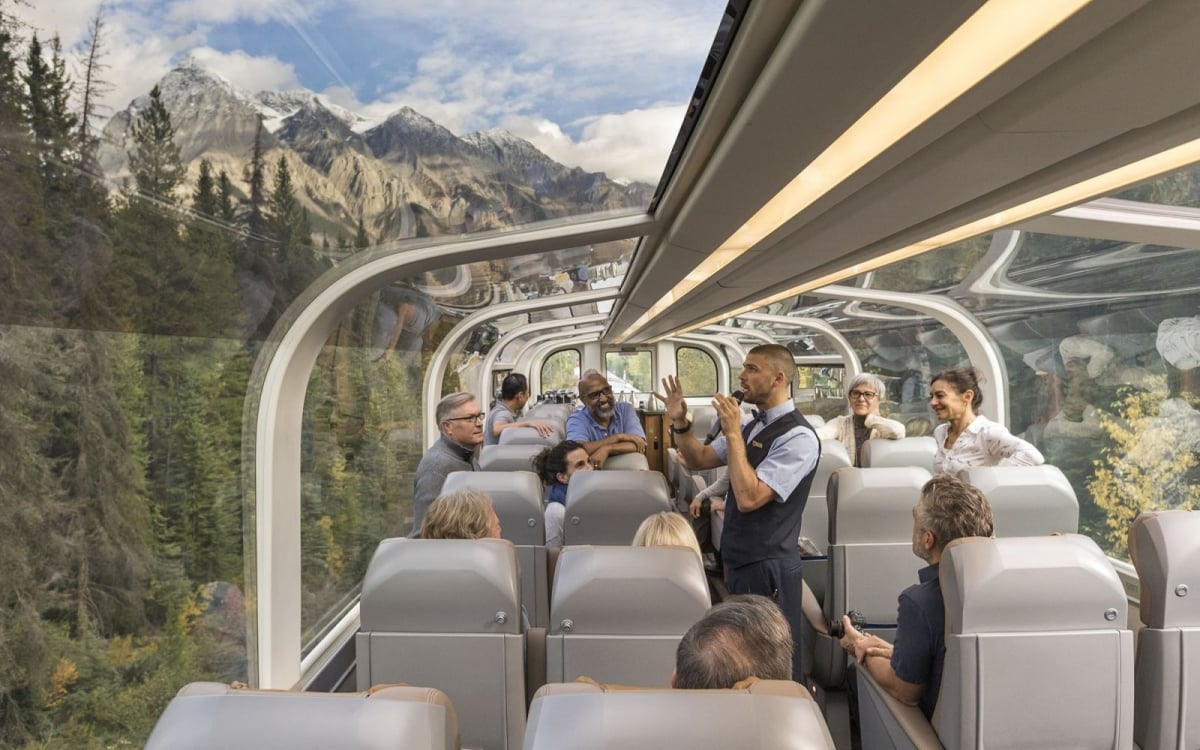 Onboard Rocky Mountaineer in Gold Leaf Train staff talk to animated passengers passing through mountain scenery