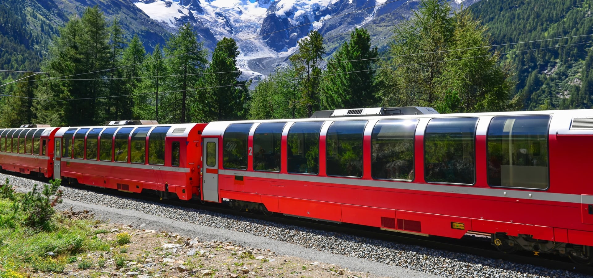 A train travels through Alpine scenery with bright blue sky