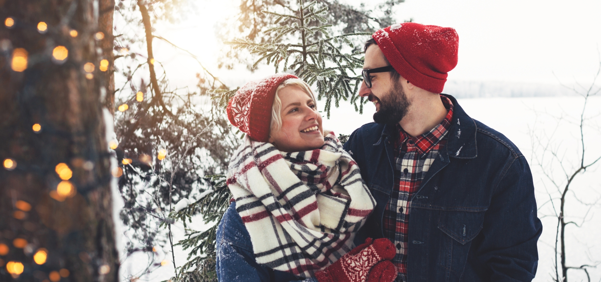 Beautiful couple in winter clothes walking along snowy forest. Woman wearing red hat, checkered scarf and mittens. Man in glasses and jean jacket