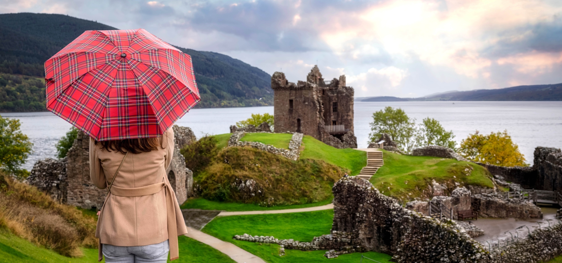 A tourist woman with a scottisch pattern umbrella looks at the famous Urquhart Castle at Loch Ness during autumn time, Scotland