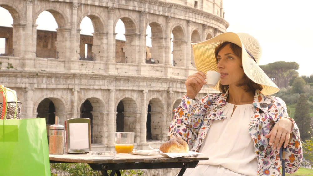 Happy young woman tourist drinking coffee and juice with cornetto at the table outside a bar restaurant in front of the Colosseum in Rome.